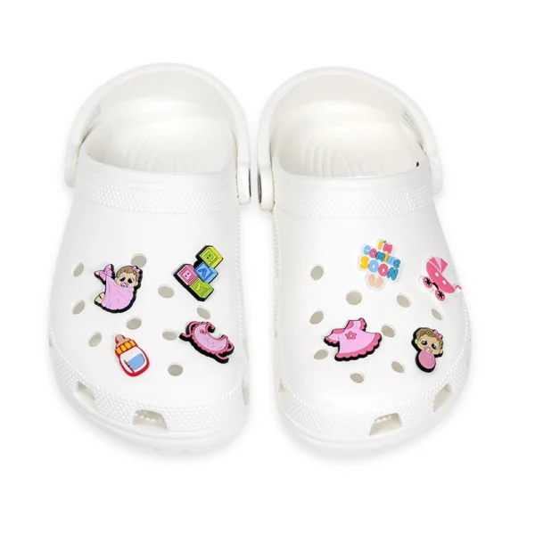 White Crocs featuring baby girl shoe charms, including a baby in a blanket, bottle, pacifier, pink dress, stroller, building blocks, and "I'm Coming Soon" charm