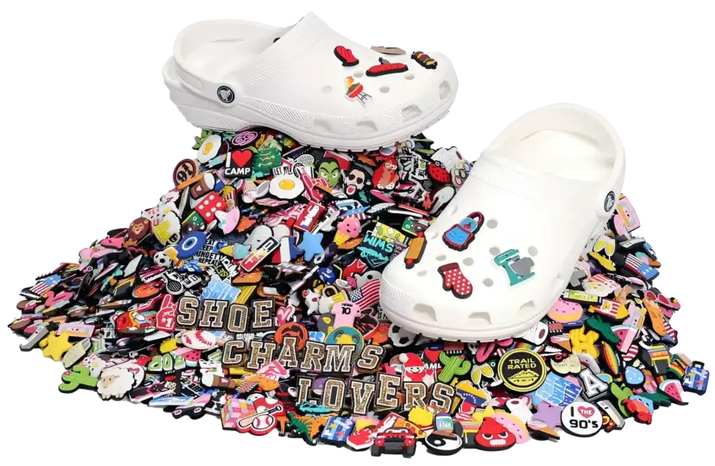 White Crocs adorned with colorful shoe charms displayed on a vibrant pile of assorted charms, featuring various themes and designs. The words 'SHOE CHARMS LOVERS' stand out among the collection, emphasizing creativity and customization