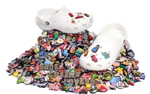 White Crocs adorned with colorful shoe charms displayed on a vibrant pile of assorted charms, featuring various themes and designs. The words 'SHOE CHARMS LOVERS' stand out among the collection, emphasizing creativity and customization